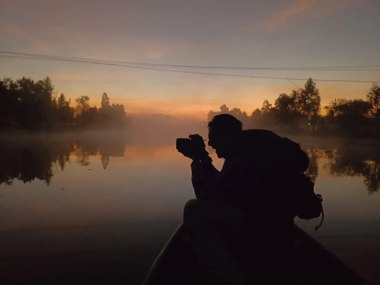 Silueta de una persona fotografiando al amanecer desde una canoa, con niebla sobre el canal y ahuejotes al fondo